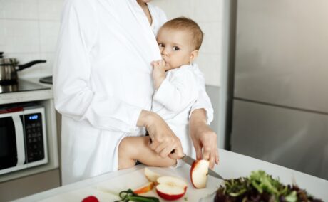 Femeie cu halat alb, care ține bebelușul, îmbrăcat în alb, cu fundul pe blatul de la bucătărie și îl alăptează în timp ce taie cu cuțitul un măr în felii