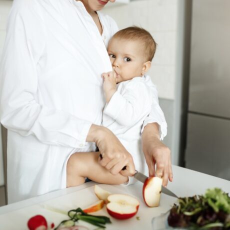 Femeie cu halat alb, care ține bebelușul, îmbrăcat în alb, cu fundul pe blatul de la bucătărie și îl alăptează în timp ce taie cu cuțitul un măr în felii