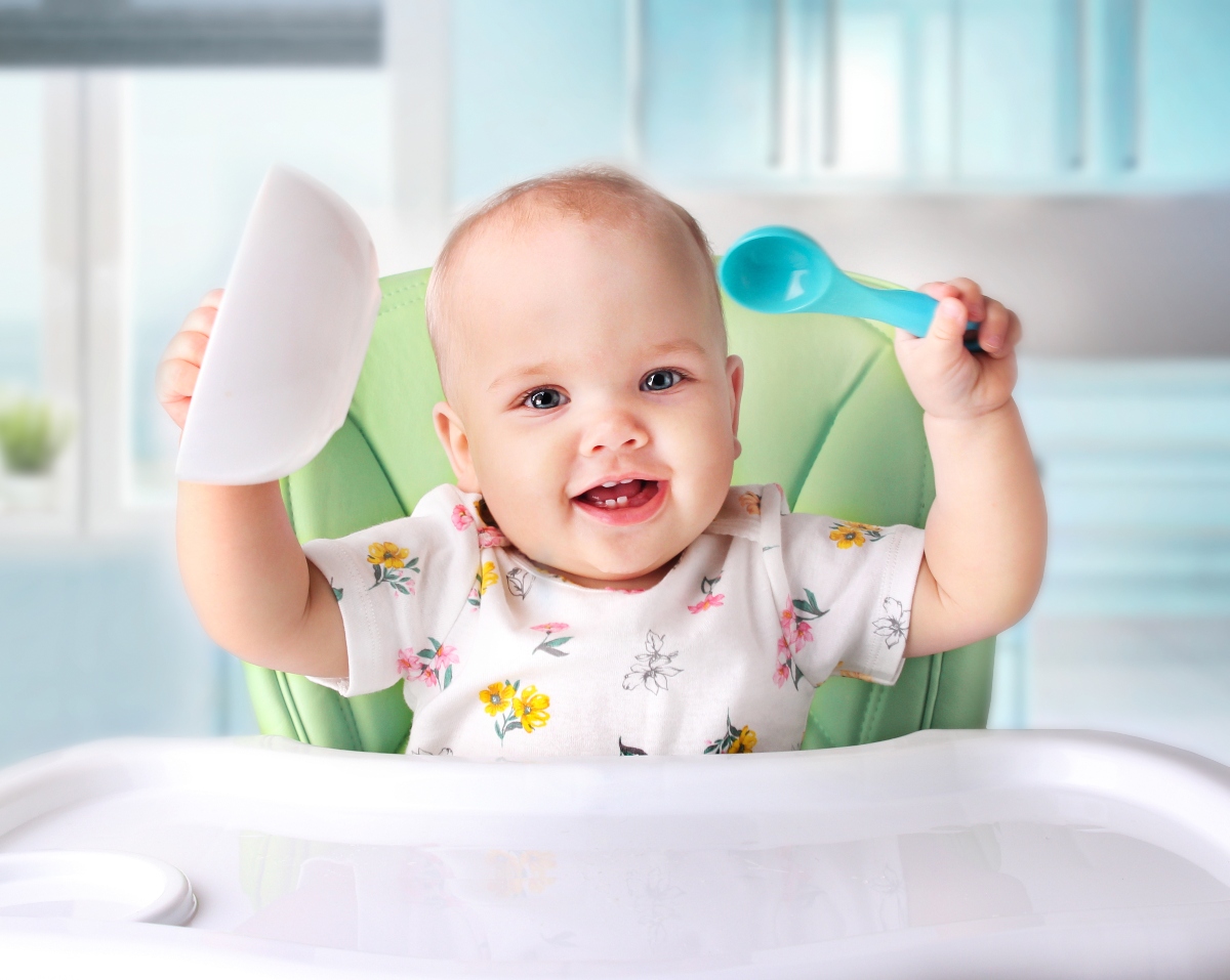 Bebeluș care stă în scaun de masă, verde, este îmbrăcat într-un body cu flori, cu mânecu scurte și ține într-o mână o lingură din plastic, albastră, și în cealaltă mână un bol alb, din plastic