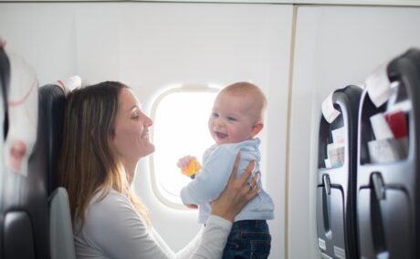 Bebeluș în avion, îmbrăcat cu o bluză bleu și jeanși, stând în picioare, pe genunchii mamei îmbrăcate cu o bluză albă