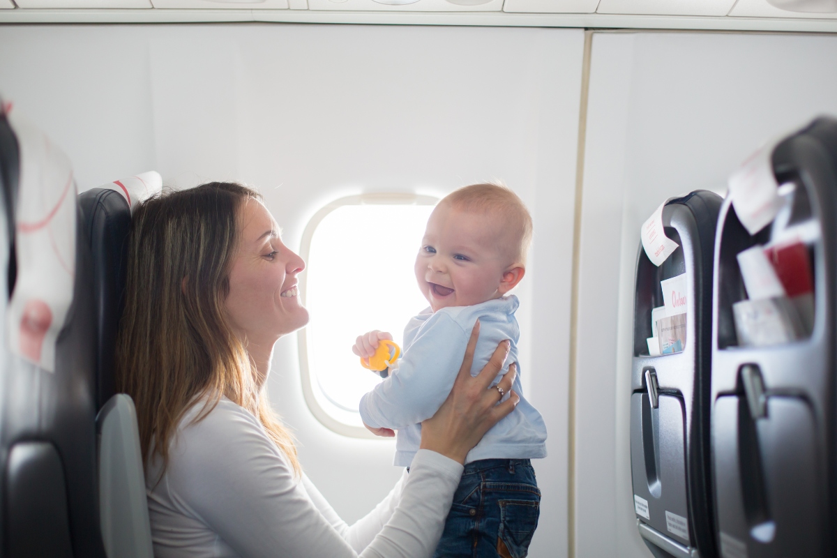 Bebeluș în avion, îmbrăcat cu o bluză bleu și jeanși, stând în picioare, pe genunchii mamei îmbrăcate cu o bluză albă