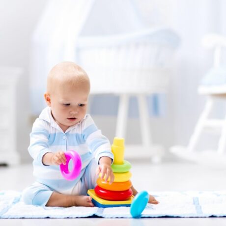 Bebeluș care stă pe jos, în camera lui, îmbrăcat într-un body cu dungi albe și bleu, și se joacă cu o jucărie stacabilă, cu cercuri colorate și ține în mână un cerc mov