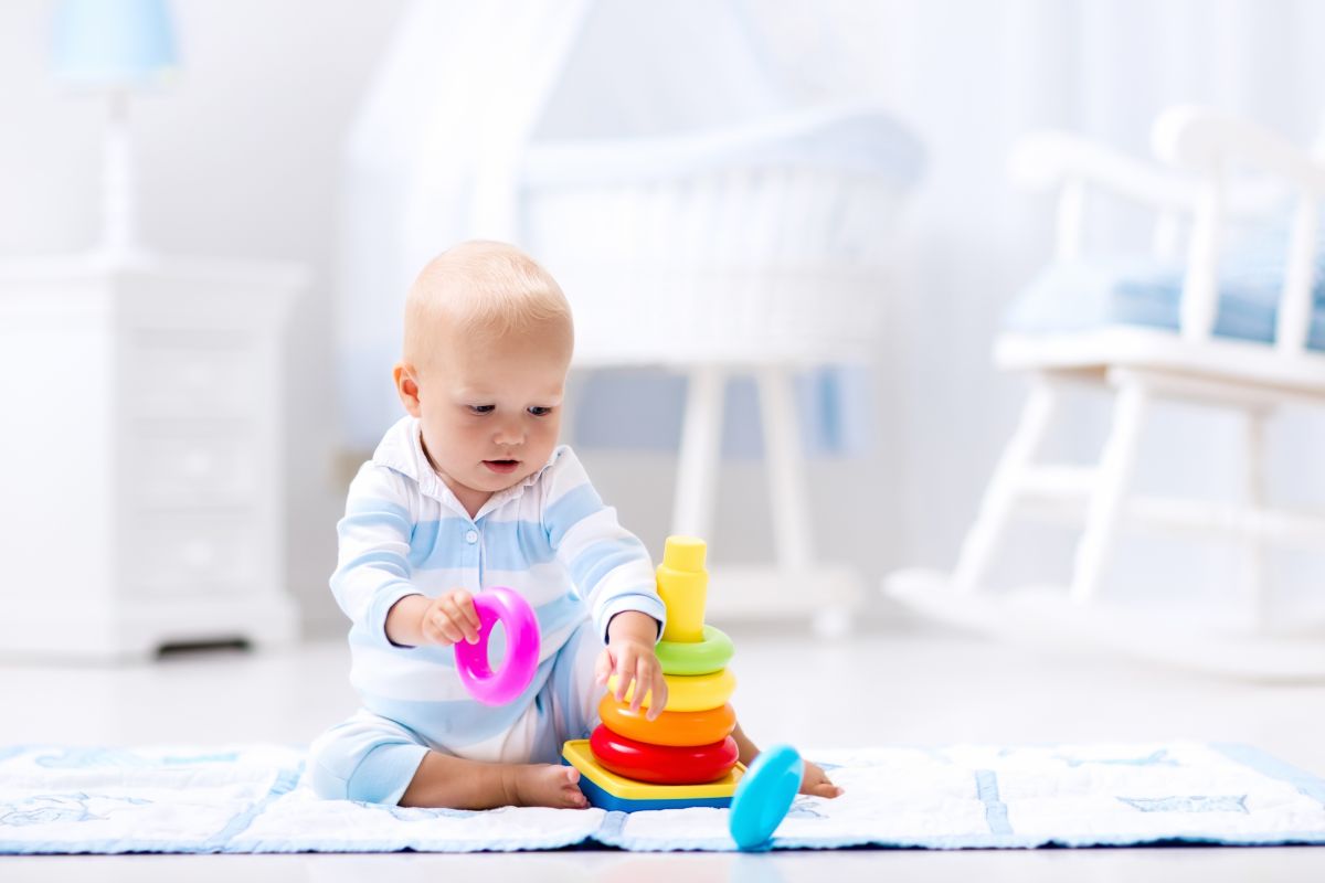 Bebeluș care stă pe jos, în camera lui, îmbrăcat într-un body cu dungi albe și bleu, și se joacă cu o jucărie stacabilă, cu cercuri colorate și ține în mână un cerc mov
