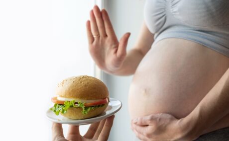 Femeie însărcinată, cu burta descoperită, care ține o mână pe burtă și cu cealaltă respinge o mână care ține pe o farfurie mică un hamburger