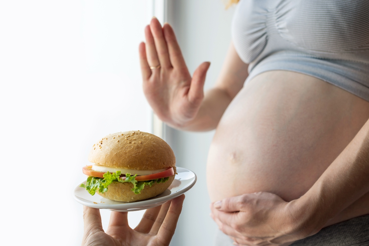 Femeie însărcinată, cu burta descoperită, care ține o mână pe burtă și cu cealaltă respinge o mână care ține pe o farfurie mică un hamburger
