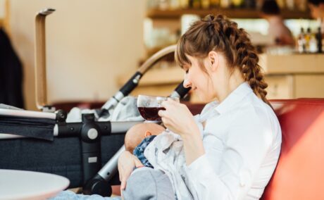 Mămică îmbrăcată cu o cămașă albă și jeanși, cu un bebeluș în brațe și o cafea în mână, lângă un căruior negru, într-o cafenea