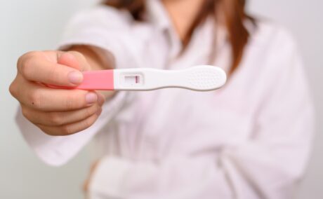 Femeie îmbrăcată în halat alb, care ține în mână un test de sarcină alb cu roz, tip stilou, negativ