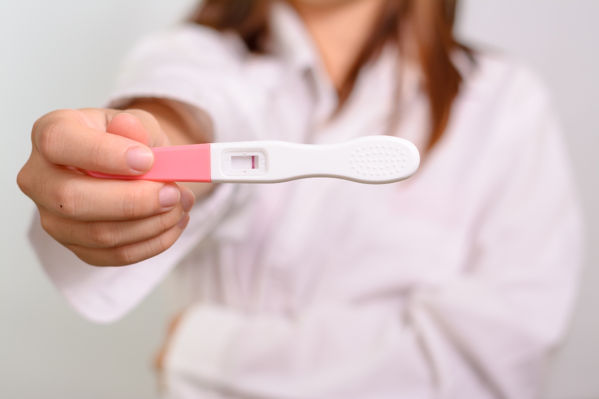Femeie îmbrăcată în halat alb, care ține în mână un test de sarcină alb cu roz, tip stilou, negativ
