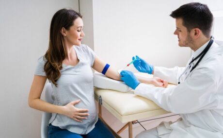 Femeie însărcinată, îmbrăcată într-un tricou gri și jeanși, care stă pe un scaun într-un cabinet medical, și un medic cu halat alb și mănuși chirurgicale, albastre, are o seringă în mână și se apropie de mâna ei să îi recolteze sânge