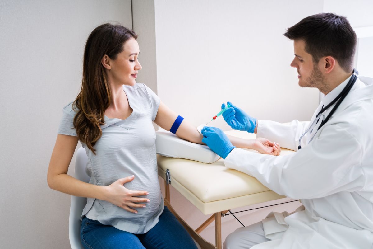 Femeie însărcinată, îmbrăcată într-un tricou gri și jeanși, care stă pe un scaun într-un cabinet medical, și un medic cu halat alb și mănuși chirurgicale, albastre, are o seringă în mână și se apropie de mâna ei să îi recolteze sânge