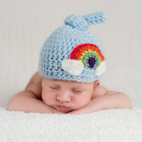 Bebeluș care doarme pe burtică cu mâinile sub bărbie și pe cap are o căciulă bleu, tricotată, cu un curcubeu pe ea