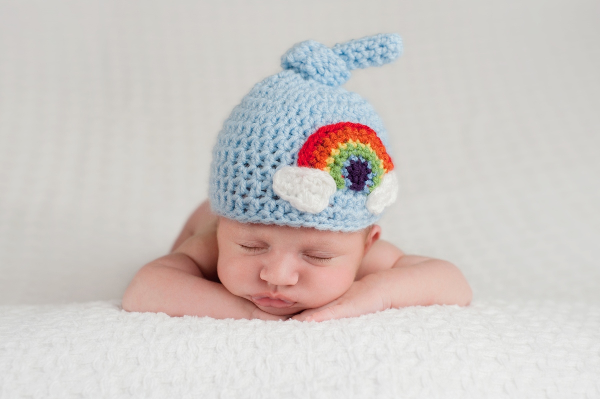 Bebeluș care doarme pe burtică cu mâinile sub bărbie și pe cap are o căciulă bleu, tricotată, cu un curcubeu pe ea