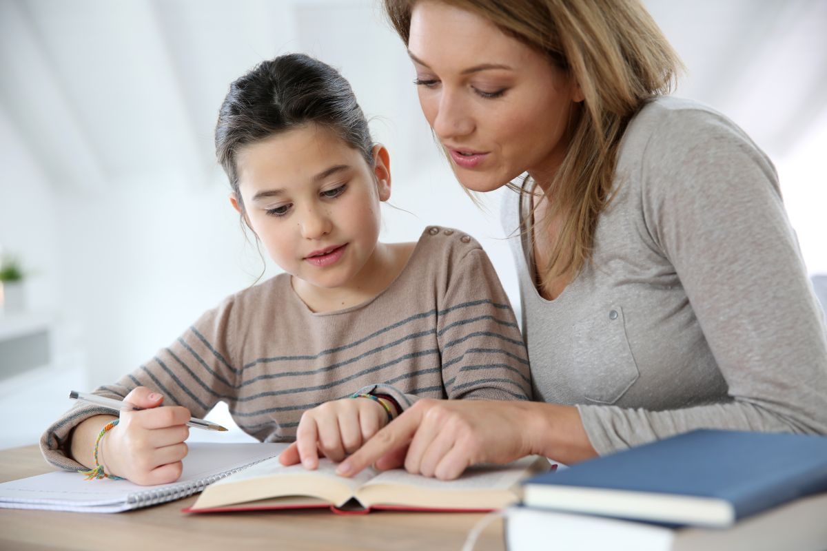 Mamă alături de fetița ei, pe care o ajută să citească, stau la un birou și fetița ține un pix în mână și cu cealaltă mână urmărește textul