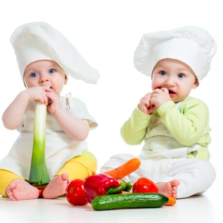 Doi bebeluși care mănâncă mai multe tipuri de legume, fotografiați pe fundal alb, cu bonete de bucătar, albe, pe cap