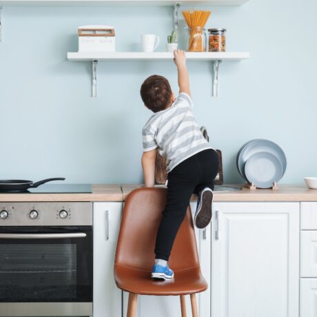 Copil în bucătătărie mobilată cu aragaz pe care stă o tigaie, dulaă și etajeră, care se urcă de pe un scaun maro, pe dulap și se agață de etajeră