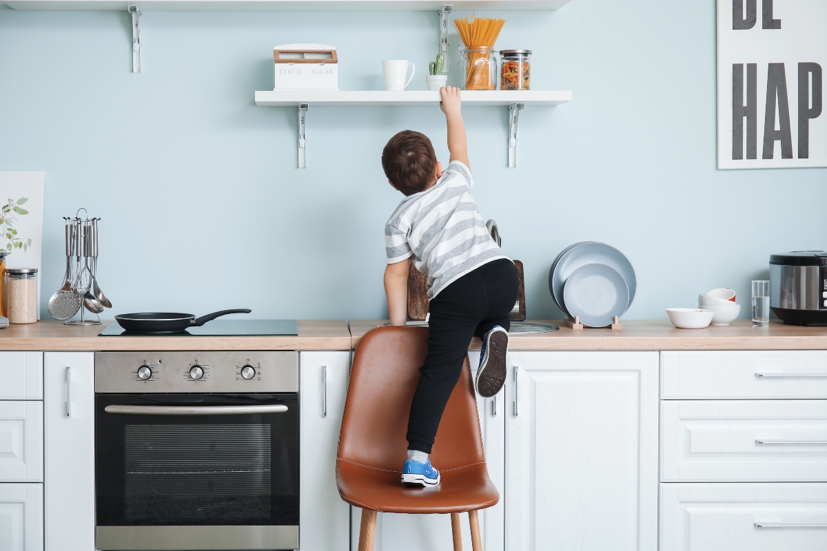 Copil în bucătătărie mobilată cu aragaz pe care stă o tigaie, dulaă și etajeră, care se urcă de pe un scaun maro, pe dulap și se agață de etajeră
