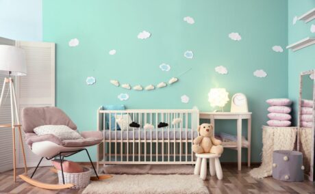 Cameră de bebeluși cu perete verde deschis și norișori aplicați, pătuț din lemn, un scaun și un scăunel pe care stău un ursuleț maro, veioză albă amre și măsuță pe care stă o veioză mică, și lângă aceasta o altă masă cu 4 perne roz