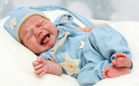 Nou-născut, care plânge așezat pe-o parte, în pat și este îmbrăcatr cu un body din tricot, bleu cu steluțe bej și nasturi aplicați și pe cap are o căciulă bleu cu steluță și nasture aplicat