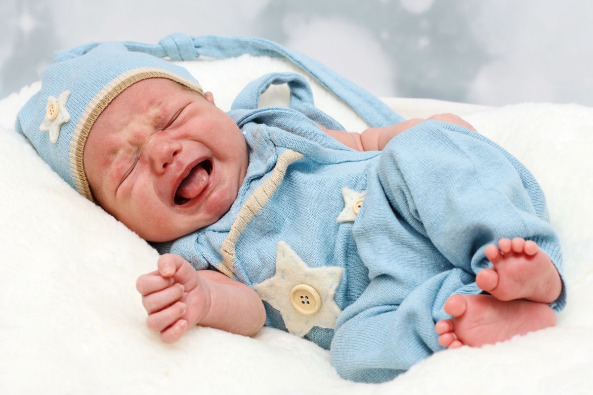 Nou-născut, care plânge așezat pe-o parte, în pat și este îmbrăcatr cu un body din tricot, bleu cu steluțe bej și nasturi aplicați și pe cap are o căciulă bleu cu steluță și nasture aplicat