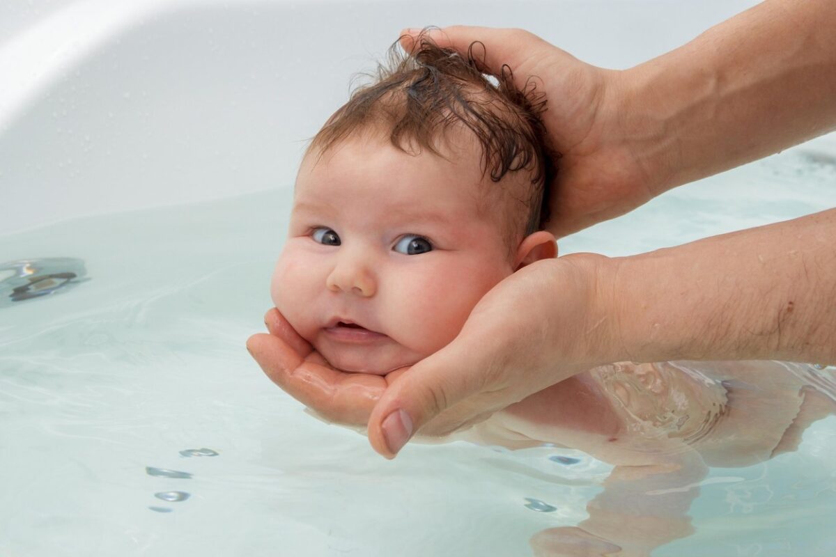 Un bebeluș care este spălat în cada de adulți și are capul sprijinit în mâinile tatălui, ilustrând de câte ori trebuie spălat bebelușul