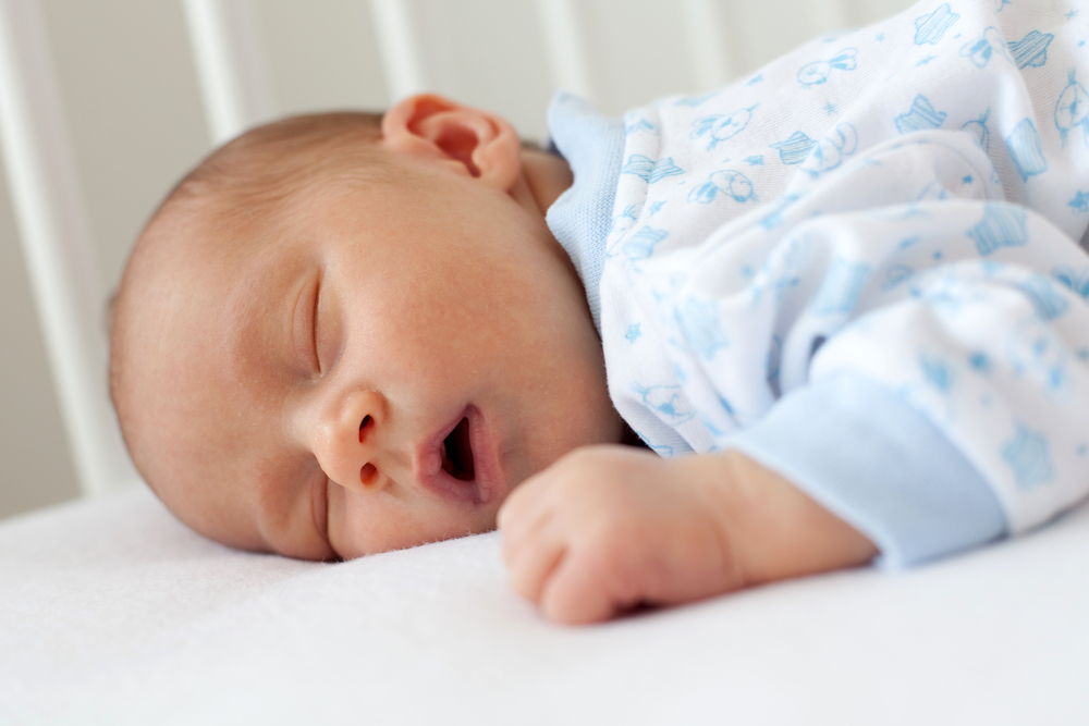 Un bebeluș băiețel, care doarme pe burtică, cu gurița deschisă, îmbrăcat într-un body alb cu imprimeu albastru