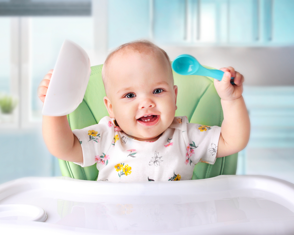 Un bebeluș care stă în scaunul de masă și ține în mâini o linguriță și un bol de mâncare