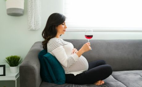 Femeie însărcinată care stă pe o canapea gri, spijinită pe o pernă verde, este îmbrăcată cu a bluză albă cu dungi negre și ține o mână pe burtă și în cealaltă mână are un pahar cu vin roșu la care se uită