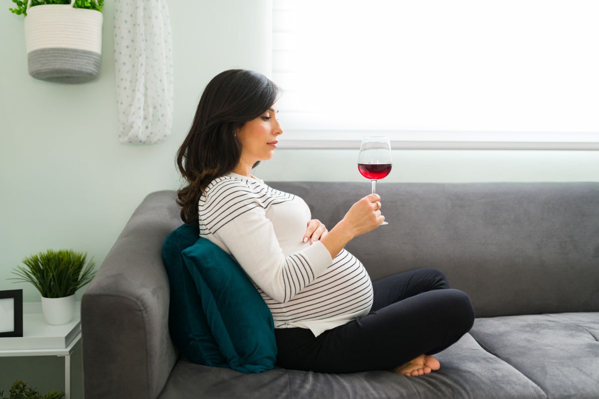Femeie însărcinată care stă pe o canapea gri, spijinită pe o pernă verde, este îmbrăcată cu a bluză albă cu dungi negre și ține o mână pe burtă și în cealaltă mână are un pahar cu vin roșu la care se uită