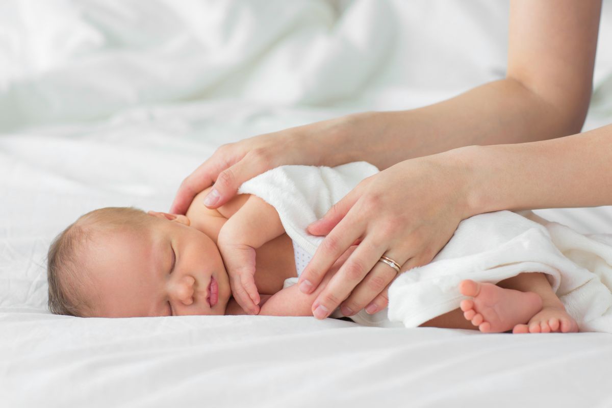 Nou-născut care doarme pe-o parte, pe un cearșaf alb, învelit cu o păturică albă și mâinile mamei peste el