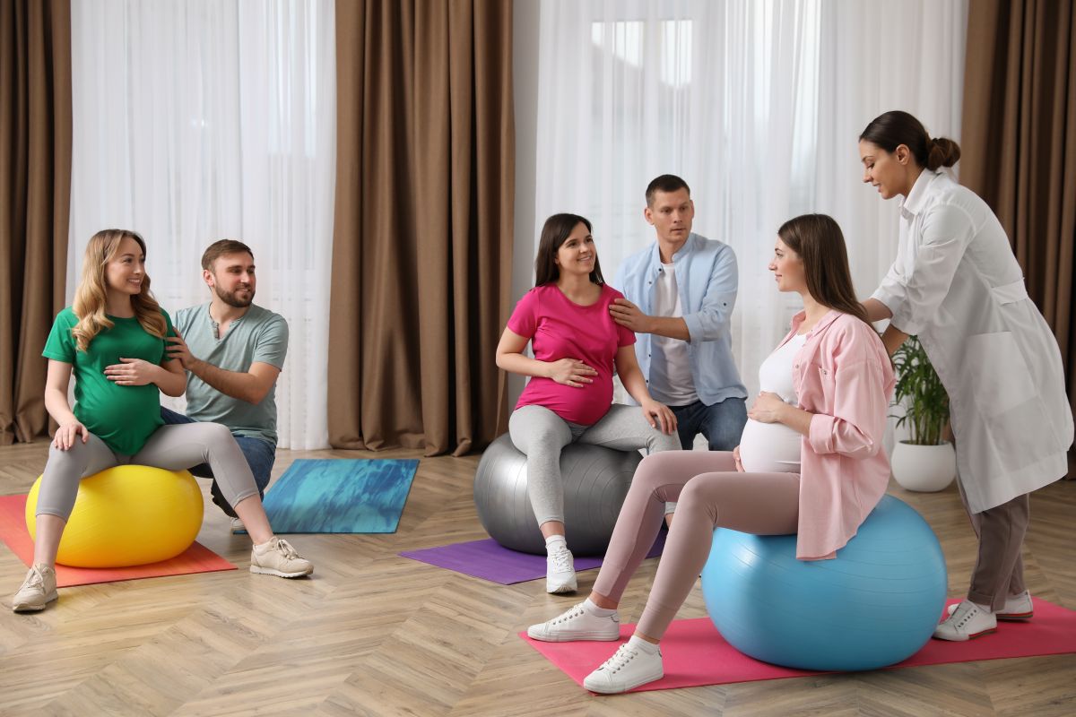 Femei însărcinate, care stau pe mingi mari, colorate, alături de partenerii lor și un specialist, la un curs la care predă metoda Lamaze