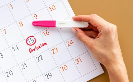 Mână de femeie, deasura unei pagini de calendar încercuite la numărul 15, care ține între degetul mare și arătător un test de ovulație alb cu roz