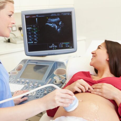 Femeie însărcinată, se uită la ecograg, stând întinsă, în cabinetul medicului ginecolog, cu o bluză roșie și burta descoperită și un doamnă doctor ginecolog care îi face o ecografie specifică morfologiei fetale pe abdomen