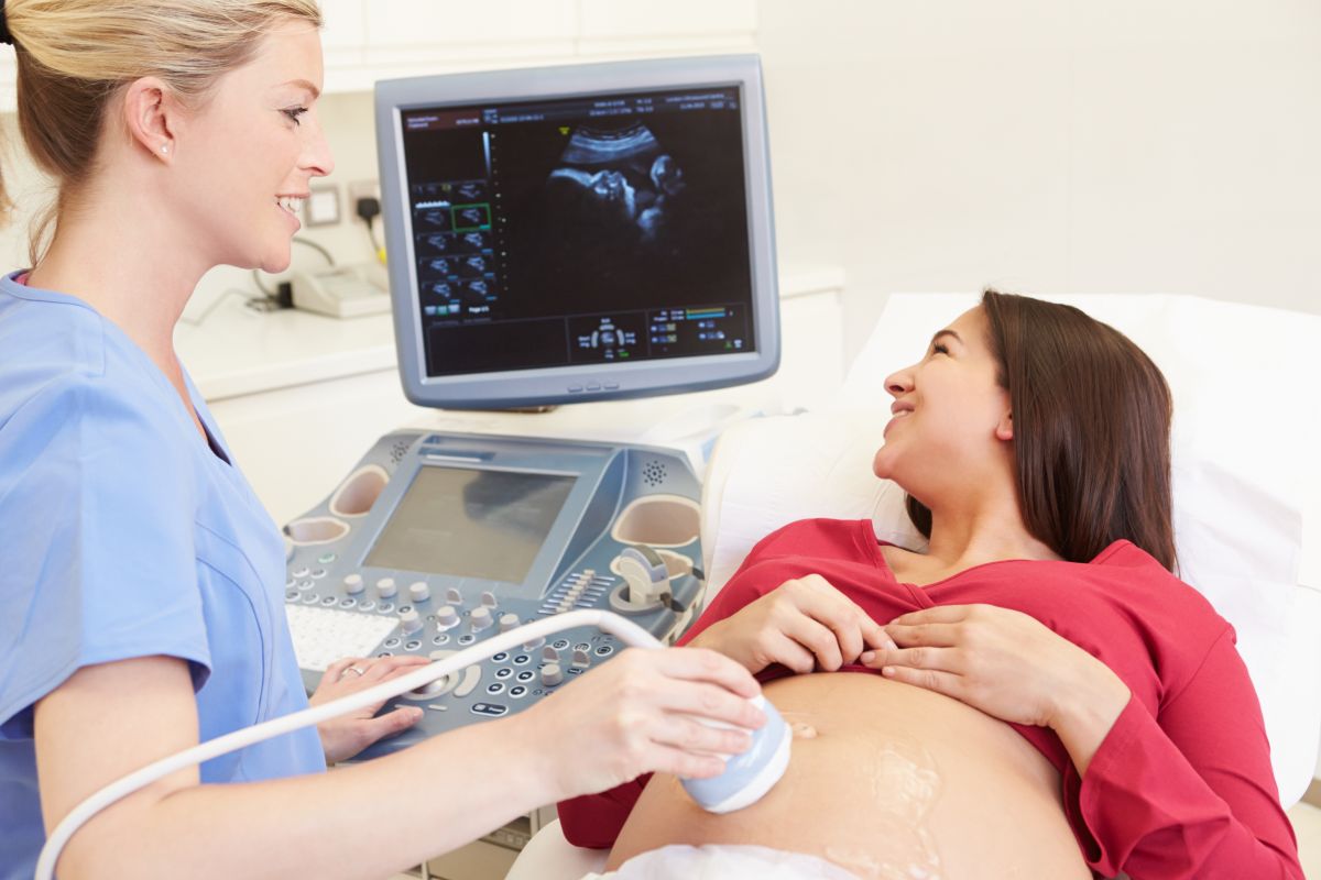 Femeie însărcinată, se uită la ecograg, stând întinsă, în cabinetul medicului ginecolog, cu o bluză roșie și burta descoperită și un doamnă doctor ginecolog care îi face o ecografie specifică morfologiei fetale pe abdomen