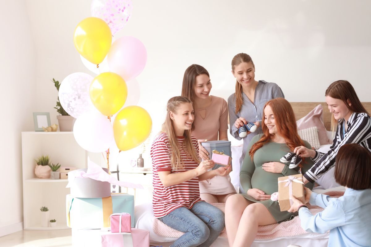 Femeie însărcinată, care stă pe canapea, în livingul decorat cu baloane colorate și cutii de cadouri și un dulap cu ghivece cu plante, alături de cinci prietene care i-au adus cadouri pentru bebeluș, ilustrând organizarea unui Baby Shower