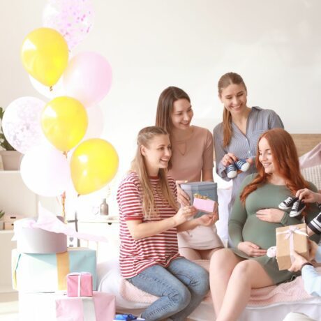 Femeie însărcinată, care stă pe canapea, în livingul decorat cu baloane colorate și cutii de cadouri și un dulap cu ghivece cu plante, alături de cinci prietene care i-au adus cadouri pentru bebeluș, ilustrând organizarea unui Baby Shower