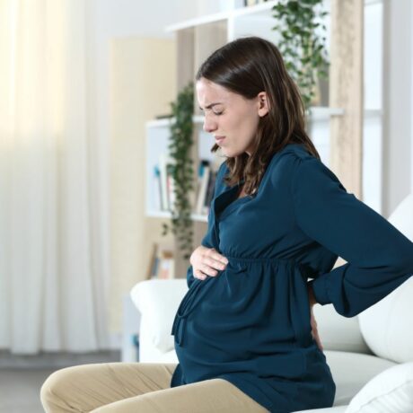 Femeie însărcinată îmbrăcată cu o bluză bleumarin și pantaloni bej stă pe o canapea albă din living și ține o mână pe burtă și una la spate