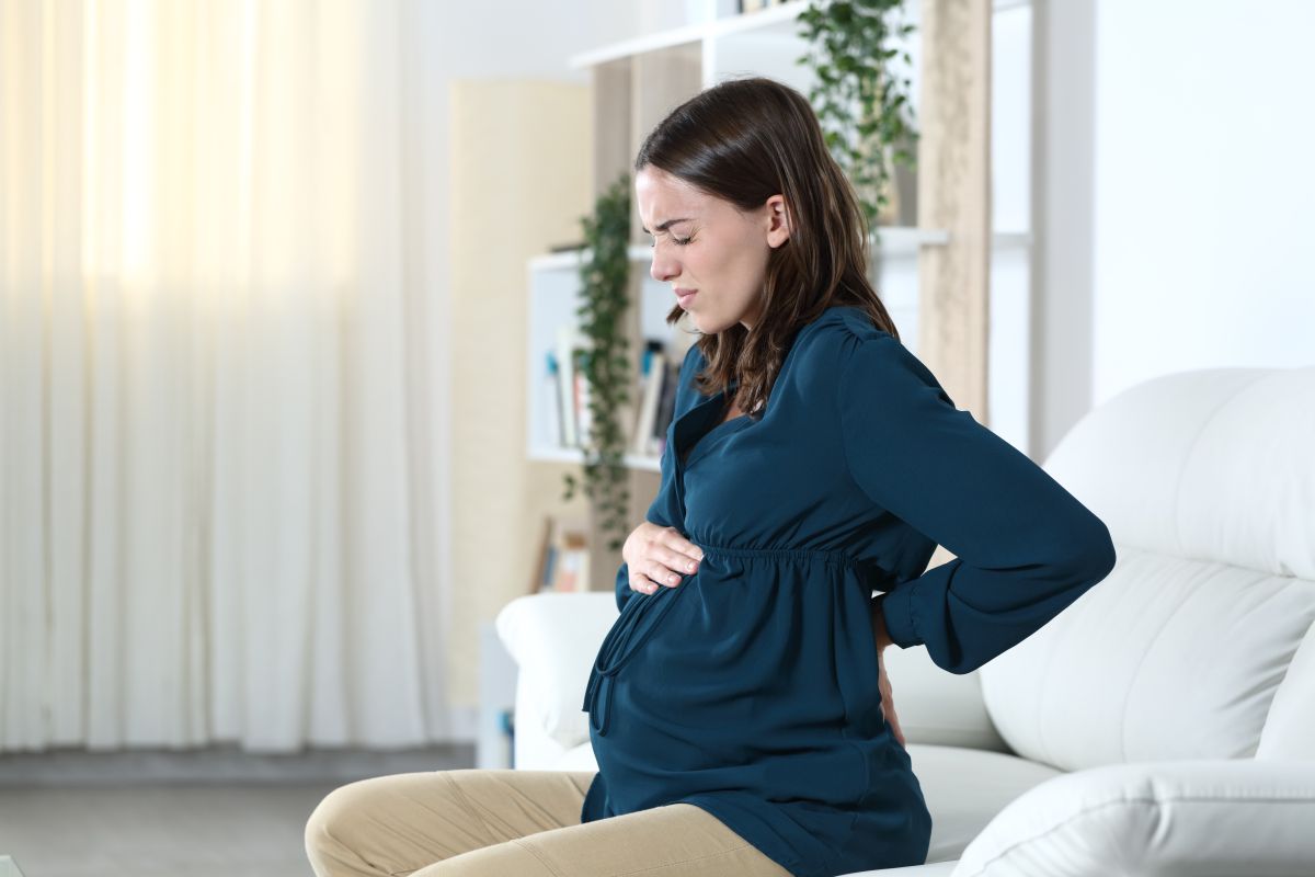 Femeie însărcinată îmbrăcată cu o bluză bleumarin și pantaloni bej stă pe o canapea albă din living și ține o mână pe burtă și una la spate