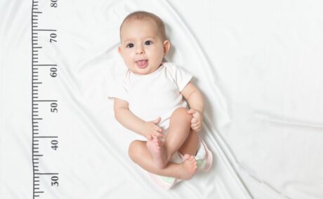 Bebeluș care stă pe spate, pe un cearșaf alb cu centimetru desenat, îmbrăcat în body alb cu mâneci scurte, cu limba scoasă, genunchii flexați și o mână pe genunchi