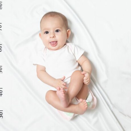 Bebeluș care stă pe spate, pe un cearșaf alb cu centimetru desenat, îmbrăcat în body alb cu mâneci scurte, cu limba scoasă, genunchii flexați și o mână pe genunchi
