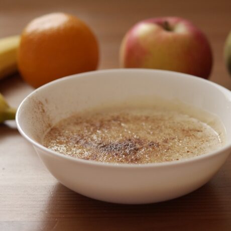 Bol alb cu porridge cu legume, așezat pe o masă din lemn, lângă o banană, o portocală, un măr și o pară