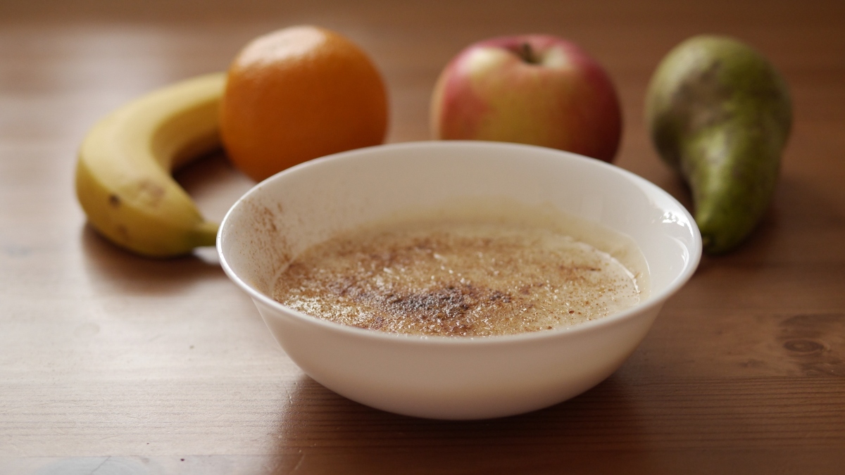 Bol alb cu porridge cu legume, așezat pe o masă din lemn, lângă o banană, o portocală, un măr și o pară