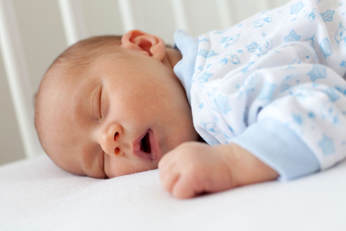 Bebeluș care doarme cu gura deschisă, în pătuț și are un body bleu cu alb, ilustrând rutina de somn a bebelușului