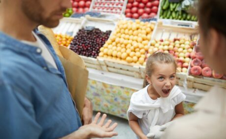 Părinți cu fetița răsfățată, care țipă, și ține în mâini o jucărie din pluș, la supermarket, lângă raionul de fructe și legume