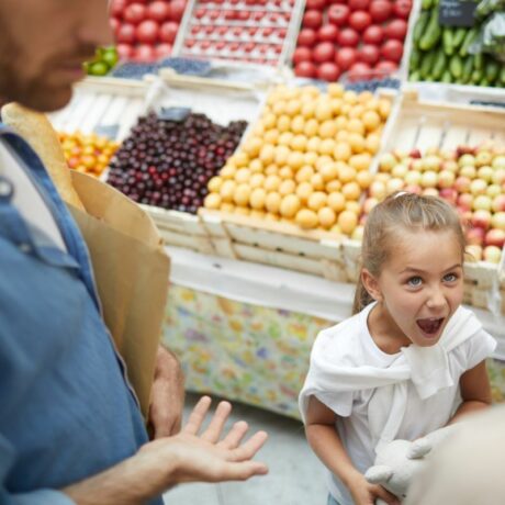 Părinți cu fetița răsfățată, care țipă, și ține în mâini o jucărie din pluș, la supermarket, lângă raionul de fructe și legume