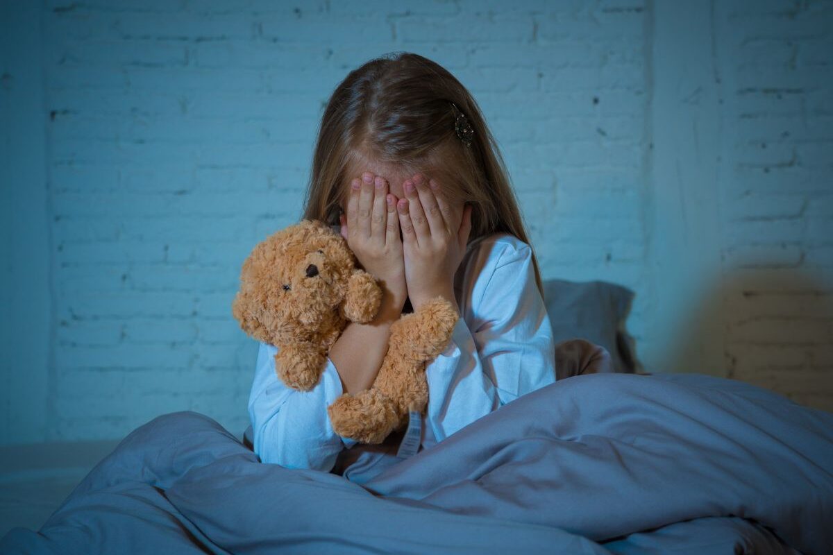 Fetiță care stă în fund, în pat, și este învelită cu o pilotă gri, ține mâinile la ochi și are un ursuleț maro în mână