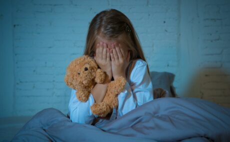 Fetiță care stă în fund, în pat, și este învelită cu o pilotă gri, ține mâinile la ochi și are un ursuleț maro în mână