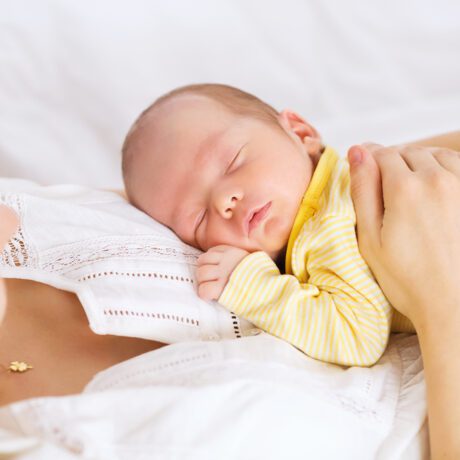 O femeie care își ține bebelușul pe piept ca să doarmă, în haine albe, în timp ce micuțul poartă un body galben