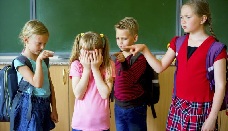 Copii la școală, care agresează o fetiță, arătând-o cu degetul și ea își acoperă fața cu mâinile