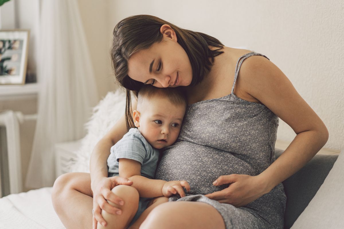 Femeie însărcinată, îmbrăcată cu o rochie gri, care stă în pat și ține o mână pe burtă și cu cealaltă ține la piept primul toddler, îmbrăcat cu un body gri, ilustrând a doua sarcină