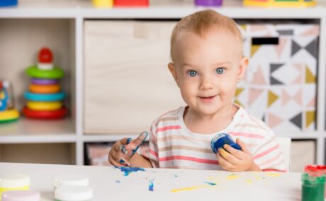 Toddler fetiță cu un tricou cu dungi colorate stă la o masă în dormitor și are în spate un dulab cu etajere pe care stau jucării, ține în mână un borcan cu vopsea albastră, pentru pictură, și este murdar de acea vopsea pe mâna cealaltă, iar masa este pictată cu vopsea albastră și galbenă și mai sunt niște borcane cu vopsea verde, roz, portocalie, ilustrând activități pe care poți să le faci cu toddlerul